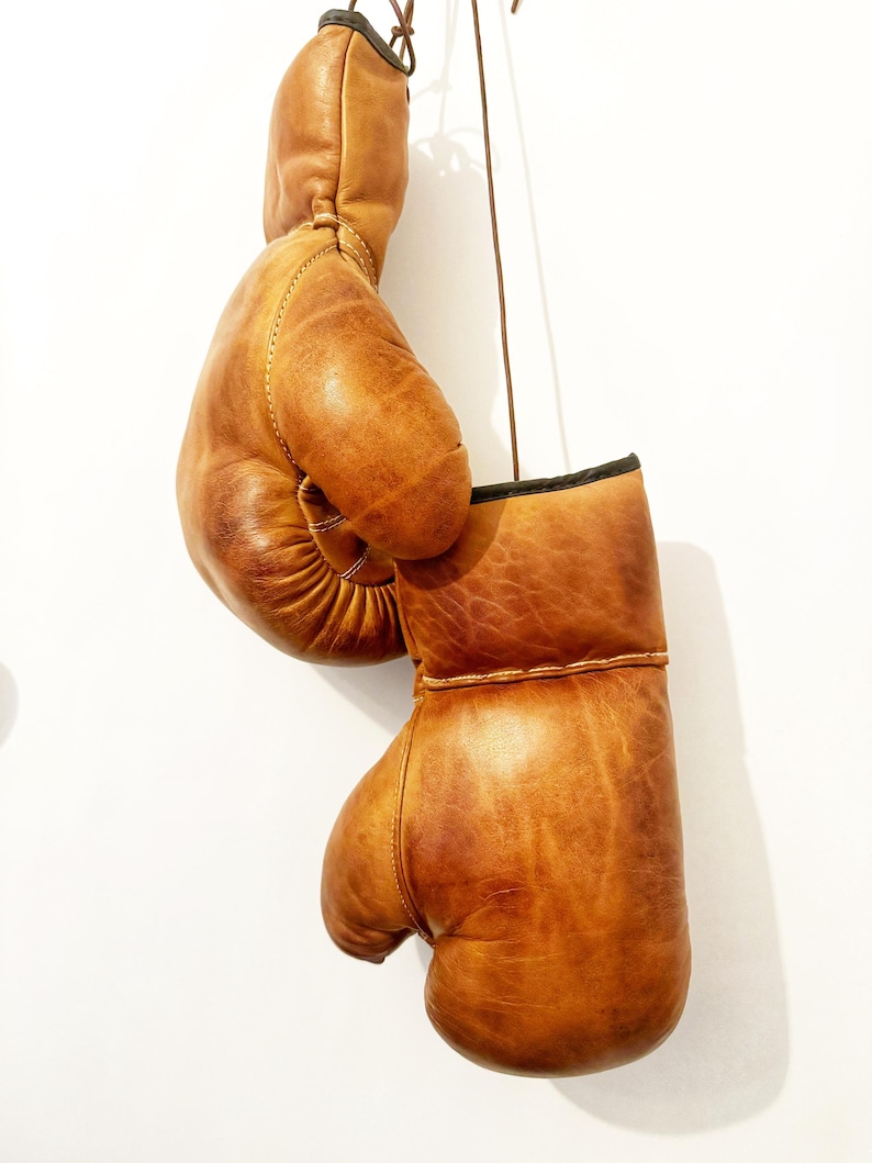 May include: A pair of brown leather boxing gloves hanging from a string. The gloves are worn and have a distressed look.