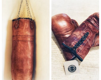 May include: A brown leather boxing bag hanging from a chain, and a pair of brown leather boxing gloves with black laces. The gloves are on a wooden surface with a small brown paper tag that says "Retro Persia" with a crossed "R" logo.
