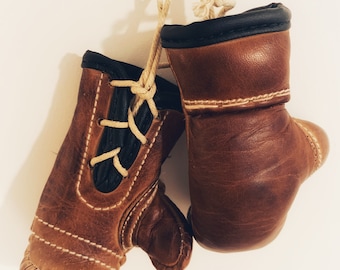 May include: A pair of brown leather boxing gloves with white stitching and black trim, hanging from a white string.