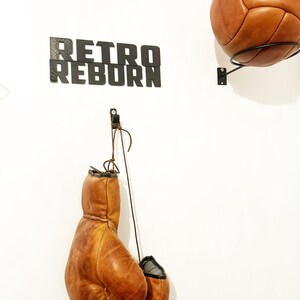May include: A white wall with a black sign that reads "Retro Reborn". There are two brown leather boxing gloves hanging from a hook and a brown leather soccer ball hanging from a hook on the wall.