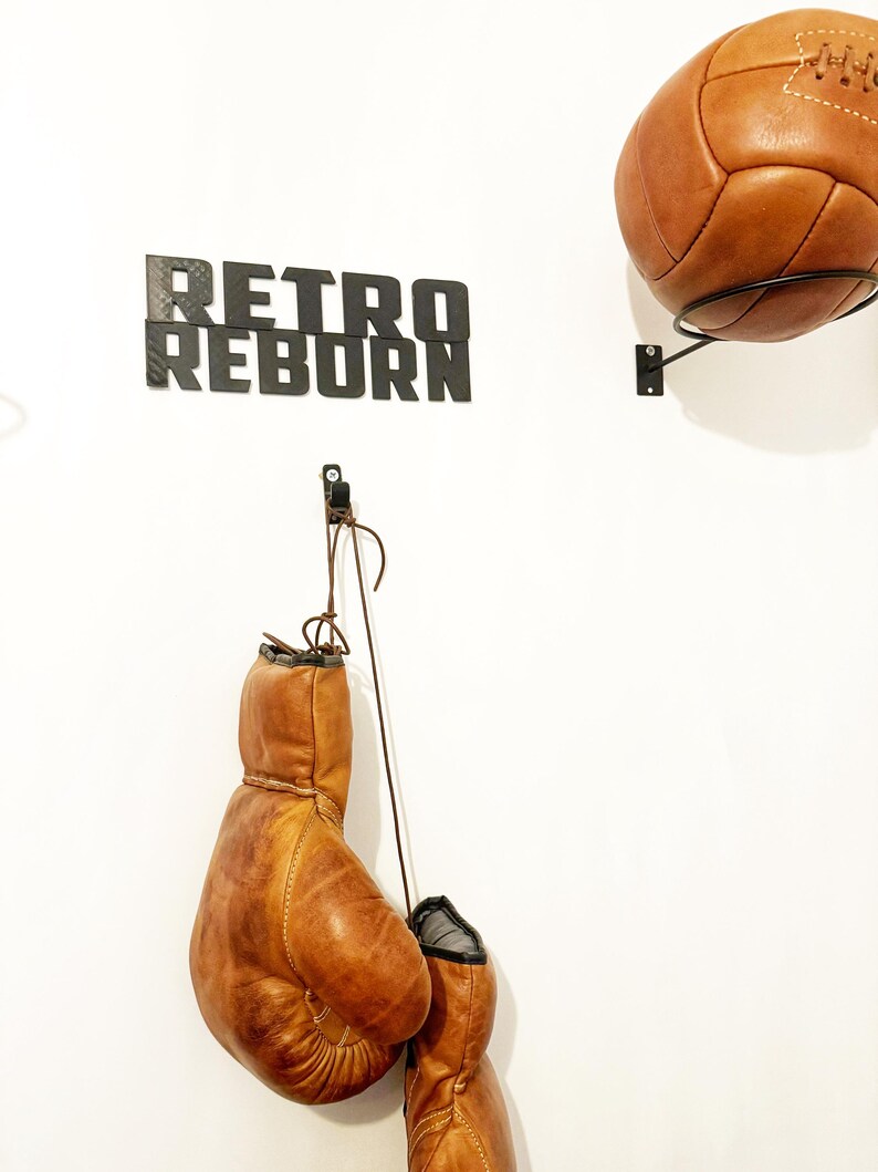 May include: A white wall with a black sign that reads "Retro Reborn". There are two brown leather boxing gloves hanging from a hook and a brown leather soccer ball hanging from a hook on the wall.