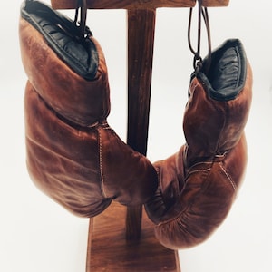 May include: A pair of brown leather boxing gloves hanging on a wooden stand. The gloves have black trim and white stitching.