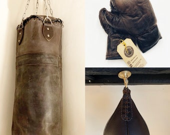 May include: Three leather punch bags hanging from the ceiling. The bags are brown and have a distressed look. The bag on the left is a traditional boxing bag, the bag in the middle is a speed bag, and the bag on the right is a heavy bag.