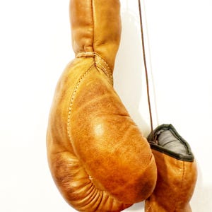 May include: A pair of brown leather boxing gloves hanging from a hook. The gloves are worn and have a vintage look.