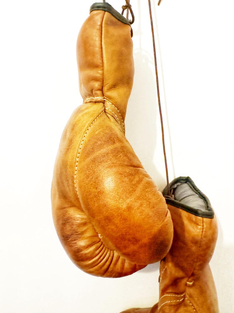 May include: A pair of brown leather boxing gloves hanging from a hook. The gloves are worn and have a vintage look.