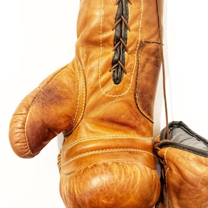 May include: A pair of brown leather boxing gloves with black laces. The gloves are hanging from a hook. The gloves are worn and have a vintage look.