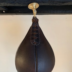May include: A dark brown leather speed bag hanging from a brass ceiling mount. The bag is teardrop-shaped with white stitching and a braided leather loop. The ceiling mount is circular and attached to a dark wooden beam.