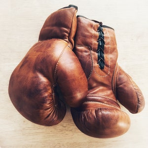 May include: A pair of brown leather boxing gloves with black laces. The gloves are worn and have a vintage look.