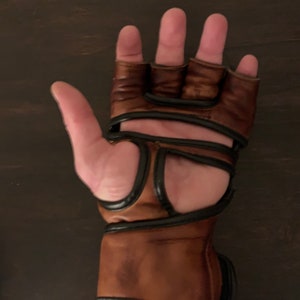 May include: A pair of brown leather fingerless gloves with black accents. The gloves have a wrist strap and are designed for training or competition.