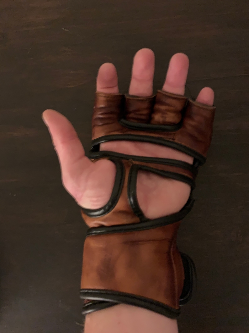 May include: A pair of brown leather fingerless gloves with black accents. The gloves have a wrist strap and are designed for training or competition.