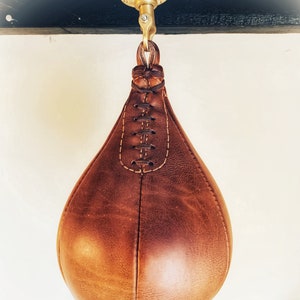 May include: A brown leather hanging punching bag with a brass hook and chain. The bag is stitched with a decorative pattern.