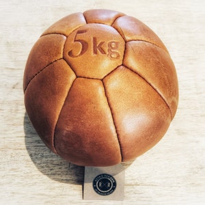 May include: A brown leather medicine ball with the text "5kg" printed on it. The ball is sitting on a light wood surface. The ball has a tag attached to it with the text "Retro Reborn" and a logo.