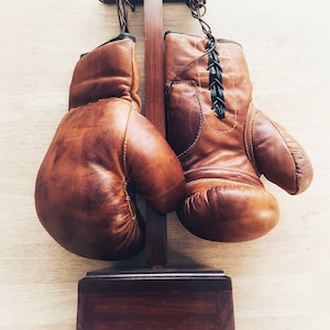 May include: A pair of brown leather boxing gloves hanging on a wooden stand. The gloves are laced with black laces and have a worn, vintage look.