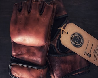 May include: A pair of brown leather fingerless gloves with black trim. The gloves are made of 100% real leather and have a distressed look. The gloves are tied together with a brown string and a tag that says "Retro Reborn" with a logo of a stylized "R" inside a circle. The tag also says "Let the old be the new" and has a social media handle and website address.