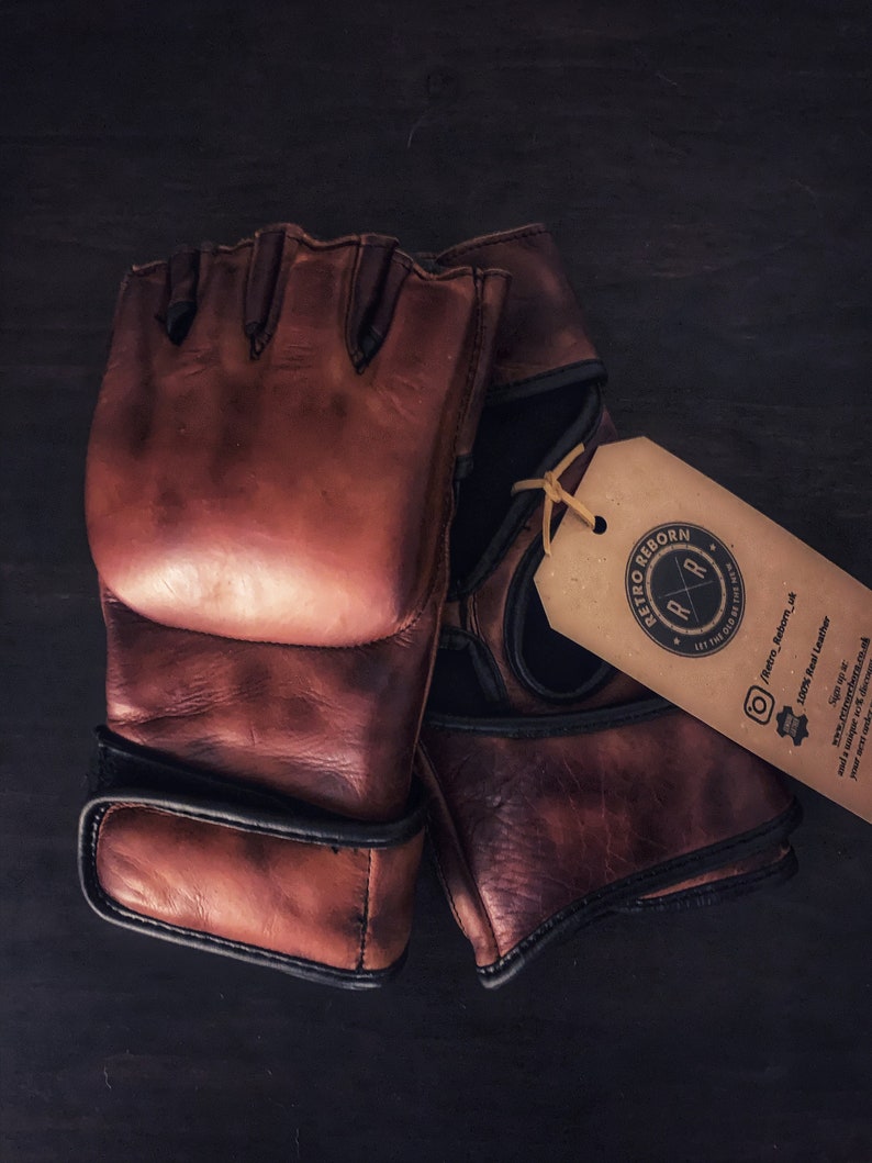 May include: A pair of brown leather fingerless gloves with black trim. The gloves are made of 100% real leather and have a distressed look. The gloves are tied together with a brown string and a tag that says "Retro Reborn" with a logo of a stylized "R" inside a circle. The tag also says "Let the old be the new" and has a social media handle and website address.