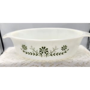 May include: A white, oval-shaped Pyrex bowl with two handles. The bowl features a floral design in dark green, with flowers and leafy sprigs. The bowl is sitting on a white, textured surface.