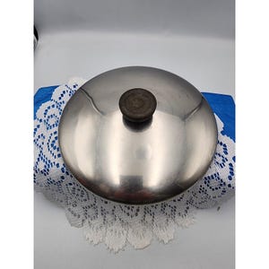 Könnte beinhalten: Ein Edelstahltopfdeckel mit einem dunkelbraunen Holzgriff. Der Deckel ist rund und reflektierend, mit einer leicht gewölbten Form. Der Deckel liegt auf einer weißen Spitzendecke und blauem Stoff.