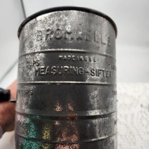 May include: Close-up of a vintage metal measuring sifter. The sifter is cylindrical with the words "BROWNELLS" and "MEASURING-SIFTER" embossed on the side. The text "MADE IN USA" is also visible. The sifter shows signs of age and wear.