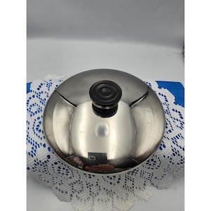 Può includere: Un coperchio per pentole in acciaio inossidabile con manico nero. Il coperchio è rotondo e riflettente, con una forma leggermente a cupola. Il manico è un pomello circolare scuro. Il coperchio è appoggiato su un centrino di pizzo bianco e un panno blu.