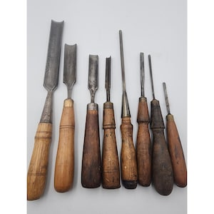 May include: A collection of seven wood carving gouges and chisels arranged in a row. Each tool features a metallic blade with a curved or flat edge and a wooden handle. The handles vary in shade and texture, showing signs of wear and age.