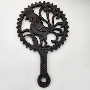 May include: Black cast iron trivet featuring a rooster design within a circular frame. The trivet has a handle for easy use and measures approximately 8 inches in diameter. The rooster is detailed with a textured finish, adding a rustic charm to the kitchen.