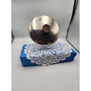 May include: A stainless steel lid with a brown knob rests on a clear plastic stand, atop a blue box covered with a white lace doily. The lid is round and reflective, with a small, circular knob.