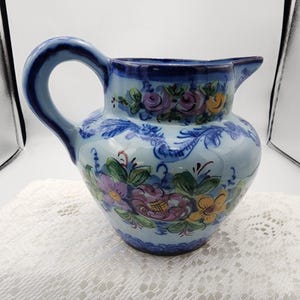 May include: A light blue ceramic jug with a curved handle and a spout. The jug is decorated with hand-painted floral designs in purple, yellow, and green. The rim and handle are a darker shade of blue.