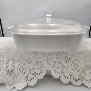 Puede incluir: Una fuente rectangular blanca con tapa de cristal transparente y un pequeño asa. La fuente tiene dos asas laterales y está sobre un mantel de encaje blanco. La tapa tiene un pequeño pomo transparente.