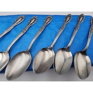May include: A set of six silver spoons with ornate floral designs on the handles. The spoons have oval bowls and are arranged on a blue surface. The spoons are made of a shiny, reflective metal.