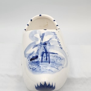 May include: White ceramic clog-shaped vase with a blue windmill design and the word "Holland" written on the side. The vase has a rounded shape with a flat bottom and is decorated with blue accents.