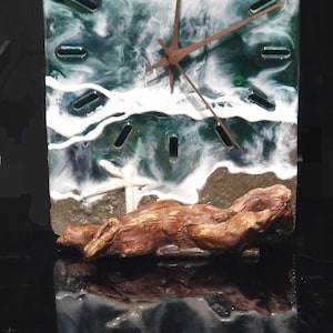 May include: A square clock with a green and white resin face depicting a beach scene. The clock hands are brown and the clock is mounted on a brown driftwood base.