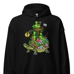 Puede incluir: Sudadera con capucha negra con un estampado gráfico de tres extraterrestres verdes montando una tortuga. Los extraterrestres llevan ropa y sostienen varios objetos, incluyendo un telescopio, un libro y una caña de pescar. La tortuga está rodeada de flores y setas.