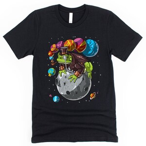 May include: A black t-shirt with a green frog wearing a top hat and monocle sitting on a moon with colorful planets in the background.