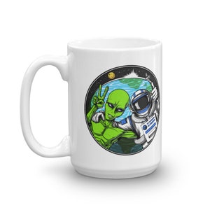 May include: White ceramic mug with a colorful graphic of an alien and an astronaut in space. The alien is giving the peace sign and the astronaut is giving a thumbs up. The graphic is in a circle with a blue and green earth in the background.