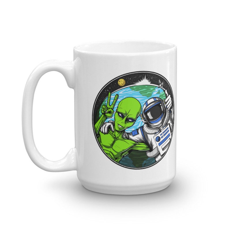 May include: White ceramic mug with a colorful graphic of an alien and an astronaut in space. The alien is giving the peace sign and the astronaut is giving a thumbs up. The graphic is in a circle with a blue and green earth in the background.