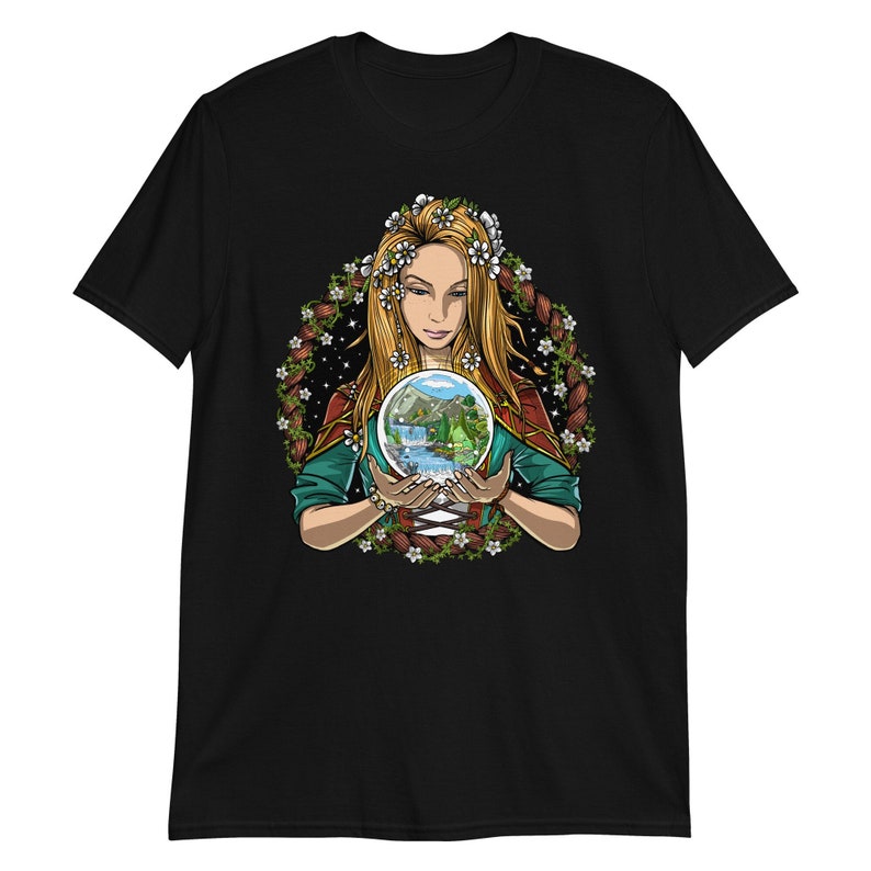 May include: Black t-shirt featuring a detailed illustration of a woman holding a crystal ball with a landscape scene inside. The woman has long blonde hair, a floral crown, and is surrounded by a floral wreath. The artwork is colorful and detailed.