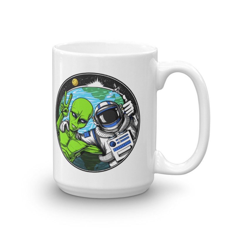 May include: White ceramic mug with a colorful illustration of an alien and an astronaut taking a selfie in front of Earth. The alien is giving the peace sign and the astronaut is giving a thumbs up.