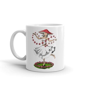 Może przedstawiać: Biały kubek ceramiczny z kolorową ilustracją kobiety w czerwono-białym kapeluszu i sukience w kształcie grzyba. Jest otoczona czerwonymi i białymi grzybami i tańczy.
