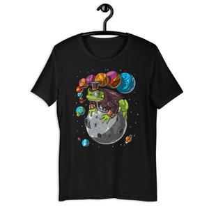 以下が含まれることがあります： 宇宙の月に座っている、シルクハットとモノクルをつけた緑色のカエルが描かれた黒いTシャツ。カエルはカラフルな惑星に囲まれている。