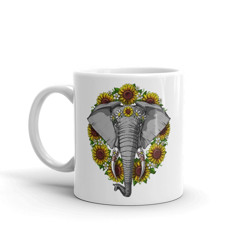 Op de afbeelding: Witte keramische mok met een grijze olifant illustratie omringd door gele zonnebloemen en witte madeliefjes. De olifant draagt een bloemenkrans.