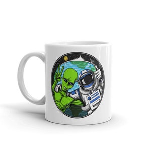May include: White ceramic mug with a colorful illustration of an alien and an astronaut giving the peace sign in front of a planet Earth. The illustration is surrounded by a circle with stars.