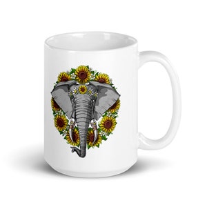 Op de afbeelding: Witte keramische mok met een afbeelding van een olifantenkop omringd door zonnebloemen en madeliefjes. De olifant draagt een bloemenkrans.