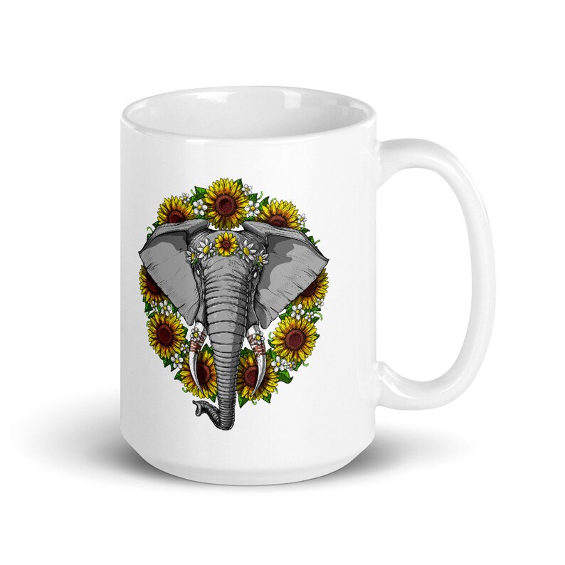 Op de afbeelding: Witte keramische mok met een afbeelding van een olifantenkop omringd door zonnebloemen en madeliefjes. De olifant draagt een bloemenkrans.