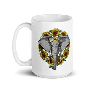 Op de afbeelding: Witte keramische mok met een afbeelding van een grijze olifant met een bloemenkrans om zijn kop, omringd door zonnebloemen. De slurf van de olifant is opgerold en zijn ogen zijn gesloten.