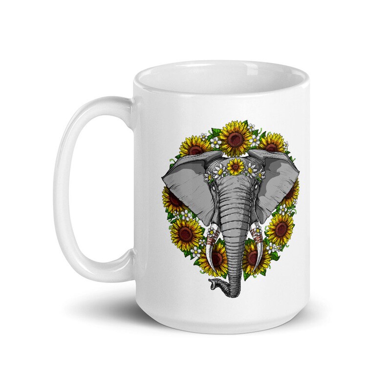Op de afbeelding: Witte keramische mok met een afbeelding van een grijze olifant met een bloemenkrans om zijn kop, omringd door zonnebloemen. De slurf van de olifant is opgerold en zijn ogen zijn gesloten.