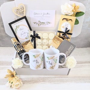 May include: A white gift box filled with wedding-themed gifts, including two mugs with "Mr." and "Mrs." on them, a framed print with the words "Olivia & Jonathan", a box of chocolates, and a bottle of body spray.