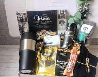Puede incluir: Una caja de regalo blanca llena de varios artículos, que incluyen una taza de viaje negra, una caja de chocolates de menta, una pastilla de jabón, una mascarilla facial, un tubo de lavado facial, un paquete de chocolate, un par de calcetines negros y una pequeña botella de spray corporal. La caja está decorada con una rama de vegetación y un trozo de cuerda.