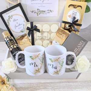 May include: A gift basket with a white background, filled with items for a wedding or engagement celebration. The basket includes two mugs with the words "Mr" and "Mrs" on them, a framed picture with a floral design, a box of chocolates, a bottle of body spray, and a box of tea lights.