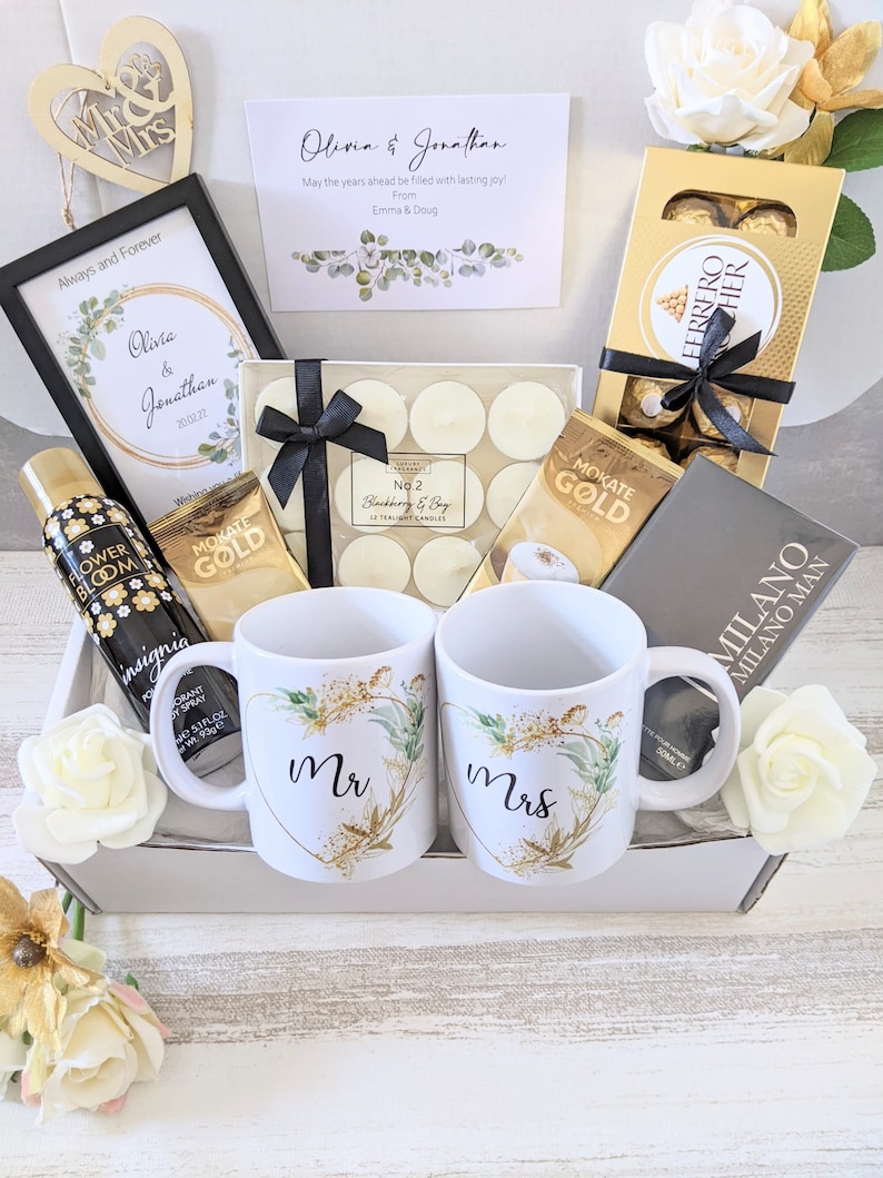 May include: A gift basket with a white background, filled with items for a wedding or engagement celebration. The basket includes two mugs with the words "Mr" and "Mrs" on them, a framed picture with a floral design, a box of chocolates, a bottle of body spray, and a box of tea lights.