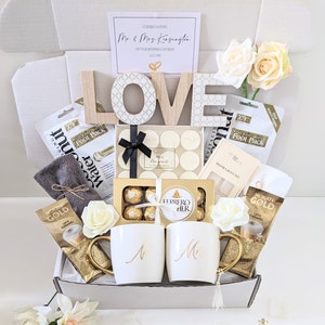 May include: A gift box filled with various items for a wedding celebration. The box includes a wooden LOVE sign, candles, chocolates, coffee, and a towel. The box is decorated with white flowers and a card that reads "Congratulations Mr & Mrs Kensington".